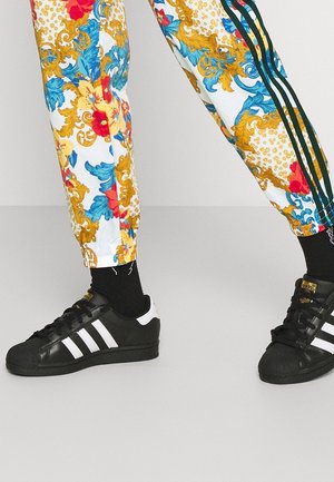 Jambes portant un pantalon blanc à motif floral avec des designs dorés et bleus et des baskets Adidas noires à rayures blanches et des chaussettes noires.