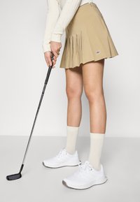 Jupe de golf plissée beige présentée avec des chaussures de sport blanches et des chaussettes beige clair, tenant un club de golf noir.