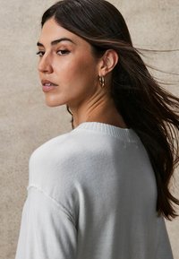 Lichtgrijze trui met geribbelde halslijn en textuurdetails, met een casual pasvorm. Het model heeft schouderlang haar en gouden ringoorbellen.