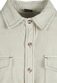 Chemise en velours côtelé beige clair avec un col pointu, trois boutons marron sur le devant et deux poches poitrine avec rabats.