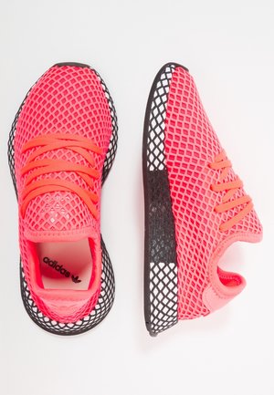 Par lyserøde Adidas sneakers med sort og hvid netmønster på sålen, lyserøde snørebånd og mesh øverste stof på hvid baggrund.