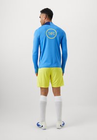 Lyseblå langærmet sportsjersey med et gult rundt logo på bagsiden, parret med klare gule shorts og hvide sokker.
