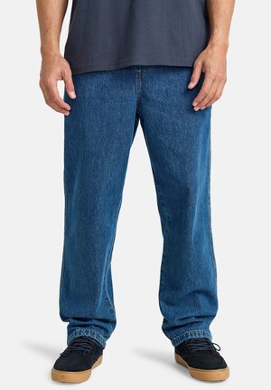 Jeans de mezclilla azul oscuro con un ajuste holgado, puños elásticos y diseño estándar de cinco bolsillos. Combinados con zapatillas negras con suelas de goma.