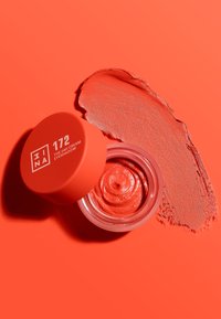 Koraalcrème oogschaduw in een doorzichtige pot met een ronde, felle koraalkleurige deksel. Zachte textuur met een swatch ernaast, alles tegen een bijpassende achtergrond.