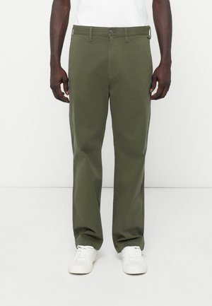 Pantalon vert olive en coton, avec une coupe droite, des poches avant et une fermeture à bouton, associé à des baskets blanches.