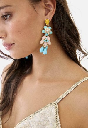 Mujer que lleva un pendiente colgante floral multicolor con piedras amarillas, azules, rosas y blancas, y una tira de camisola texturizada en crema.