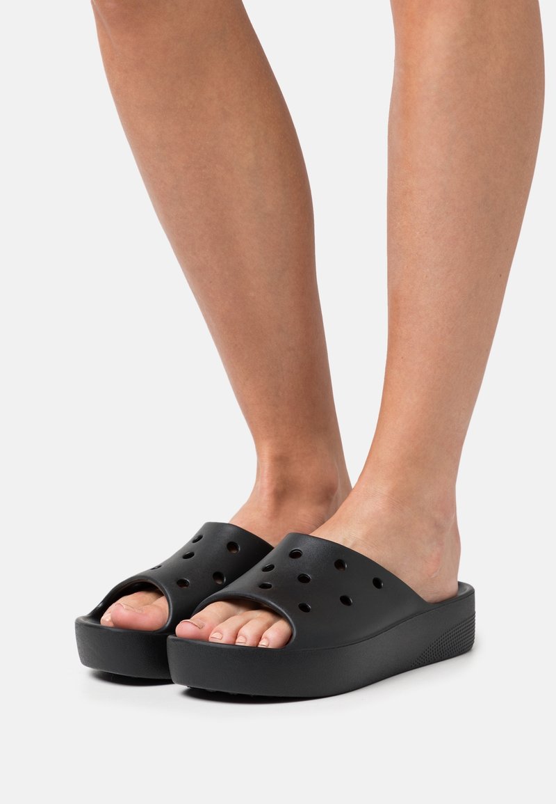 Jambes portant des sandales slides noires à plateforme avec orteils ouverts et multiples trous de ventilation sur un fond clair.
