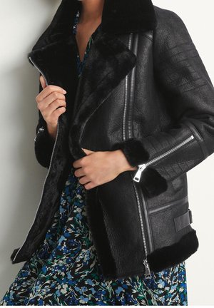 Personne portant une veste en shearling noire avec des détails de fermeture éclair sur une robe fleurie bleue et verte, tenant le col de la veste ouvert.