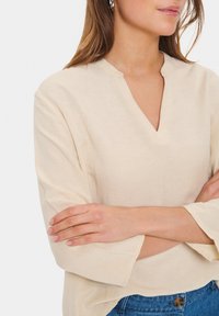 Blusa beige con scollatura a V, vestibilità ampia e maniche a campana a tre quarti, realizzata in un tessuto leggero e testurizzato.