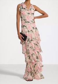 Floral print dress in soft pink and cream, featuring a tiered ruffle design, single shoulder bow accent, and a V-neckline. Complemented by a black clutch.
