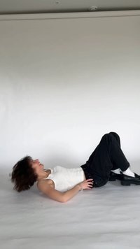 Calças de ganga pretas, top branco de alças e sapatos de plataforma pretos. A pessoa está deitada numa superfície clara, com as mãos na anca e cabelo despenteado.