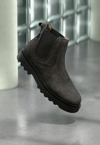 Σκούρο γκρι σουέντ μποτάκι Chelsea με μαύρα ελαστικά πλαϊνά πάνελ και ανθεκτική σόλα, κρεμασμένο πάνω από ένα γυαλιστερό δάπεδο κοντά σε τοίχο από γυάλινα τούβλα.