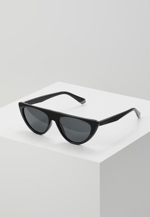 Occhiali da sole cat-eye neri con lenti scure appoggiati su un cubo bianco, con il marchio Polaroid visibile sulla parte laterale.