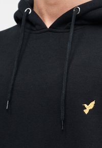 Μαύρο φούτερ με κουκούλα με πλεκτά κορδόνια, ασημένιες γκρόμεττες και μικρό κίτρινο κεντητό λογότυπο πουλιού στο αριστερό στήθος. Μαλακή υφή υλικού.