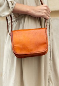Borsa a tracolla in pelle marrone con pattina, cuciture ordinate e tracolla regolabile, tenuta da una persona che indossa un vestito beige.