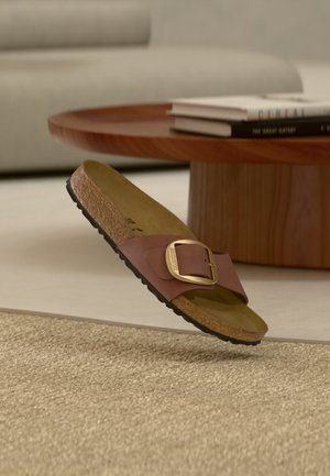Καφέ σαγιονάρα με μεγάλο χρυσό πόρπη, φελλό σόλα και μαύρη πάτημα, που βρίσκεται πάνω σε μια υφασμάτινη μπεζ μοκέτα κοντά σε ξύλινο τραπέζι με βιβλία.