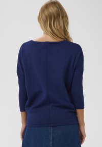Pull en maille bleu marine à coupe décontractée, avec des manches trois-quarts, un col large et une couture centrale au dos. Texture du tissu légère.