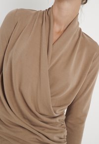 Beige Drapes mit langen Ärmeln aus weichem Stoff, mit einem Schalkragen und lockerem Schnitt, der entlang des Körpers geraffte Textur aufweist.