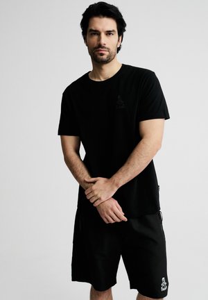 Mężczyzna o ciemnych włosach i brodzie, ubrany w czarny t-shirt i czarne szorty, stojący z rękami splecionymi z przodu na jednolitym tle.