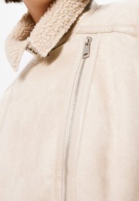 Beige jas met een pluizige, gestructureerde kraag en een diagonale ritspocket. Gemaakt van een zachte stof, met een strak en modern ontwerp.