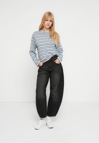 Μπλε και λευκό ριγέ πουκάμισο με μακριά μανίκια συνδυασμένο με άνετο μαύρο τζιν και λευκά αθλητικά παπούτσια. Το μοντέλο στέκεται μπροστά σε απλό λευκό φόντο.