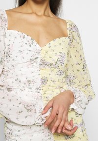 Vestido floral con escote cuadrado, confeccionado en una suave tela blanca y amarilla, con detalles fruncidos y mangas largas abullonadas con puños elásticos.