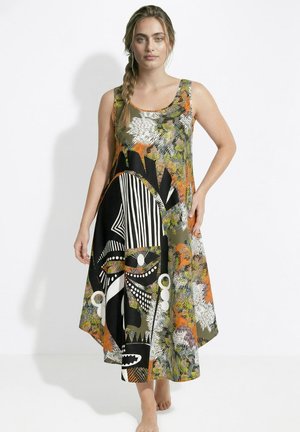 Nainen, jolla on punotut hiukset, pukeutunut hiukseton värikkääseen midimekkoon, jossa on abstrakteja mustia, valkoisia, oransseja ja vihreitä kuvioita, seisoo paljain jaloin.