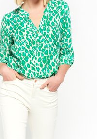 Green and turquoise leopard-print blouse with a placket and collar, long sleeves rolled to the elbows, worn with white pants.