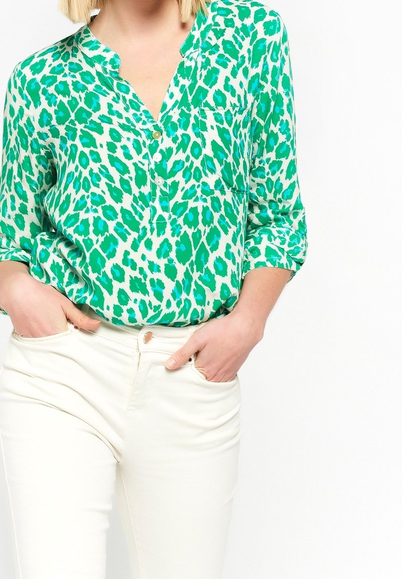 Green and turquoise leopard-print blouse with a placket and collar, long sleeves rolled to the elbows, worn with white pants.