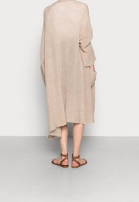 Cardigan in maglina beige, stile kimono, con maniche lunghe e ampie, scollatura aperta, due tasche laterali e orlo asimmetrico. Indossato con sandali marroni.