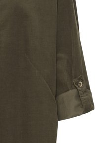 Gros plan d'une manche de chemise en velours côtelé vert olive, retroussée avec un poignet boutonné visible sur un fond uni.