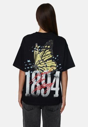 Persoon met lang, donker haar draagt een zwart oversized t-shirt met een gele vlinder, blauwe sterren, een grote witte "1804" en rood scripttekst op de achterkant.