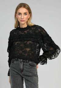 Blouse noire transparente avec des détails en dentelle florale, manches volantées et col montant. Associée à un jean gris taille haute.