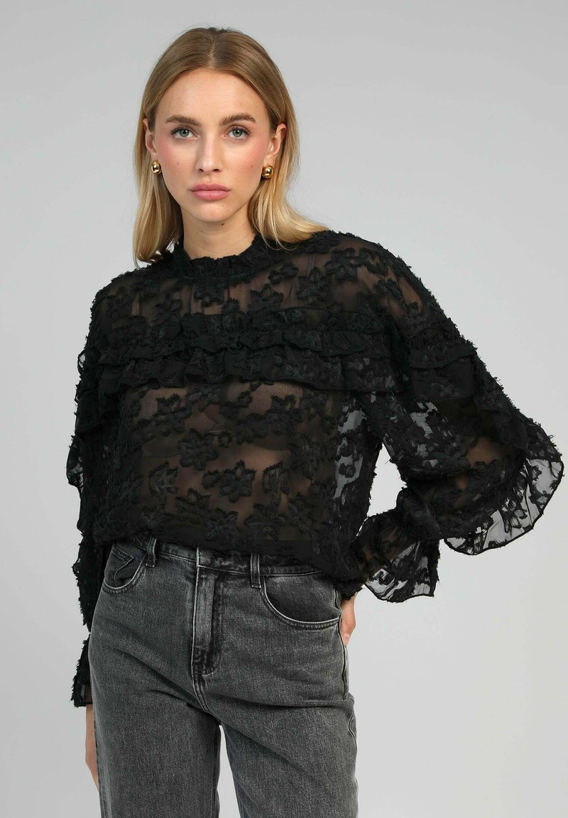 Blouse noire transparente avec des détails en dentelle florale, manches volantées et col montant. Associée à un jean gris taille haute.