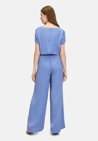 Combinaison bleu clair avec manches courtes retroussées, design à jambes larges, taille élastique, texture lisse et détail de fermeture dans le dos.