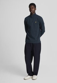 Maglia navy a maniche lunghe con zip realizzata in tessuto morbido, con un logo giallo sul petto. Indossata con pantaloni neri larghi e sneaker bianche.