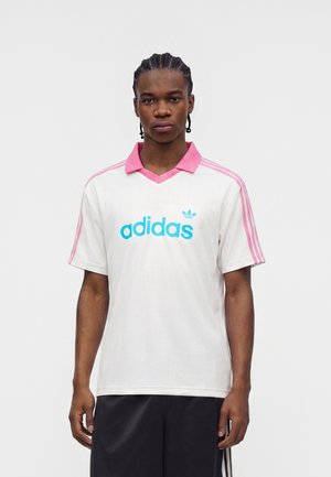 Nuori mies, jolla on letitetyt hiukset, pukeutuneena valkoiseen Adidas-paitaan, jossa on vaaleanpunainen kaulus ja raidat, sininen logo sekä mustat shortsit, seisoo yksiväristä taustaa vasten.