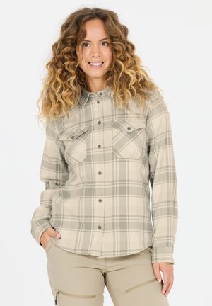 Lächelnde Frau mit lockigem Haar, die ein beiges kariertes Hemd mit Knöpfen und helle, beige Hosen mit Reißverschlusstaschen trägt, steht mit einer Hand in der Tasche.