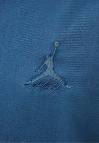 Tessuto blu con la silhouette ricamata di colore blu scuro di un giocatore di basket che salta, in possesso di una palla, con testo stilizzato sottostante.