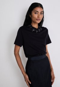 Zwarte top met korte mouwen en een gestructureerde kraag versierd met metalen accenten, gecombineerd met een donkere, ingewikkeld geweven rok.