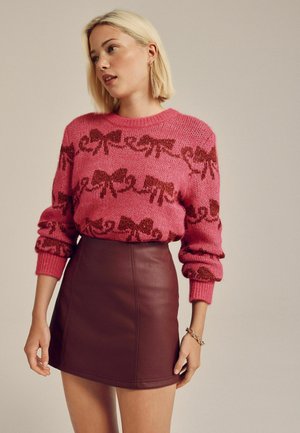 Rosa gestrickter Pullover mit rotem Schleifenmuster, gerippte Bündchen, kombiniert mit einem bordeauxroten Leder-Minirock mit klaren Linien und subtilen Nähten.
