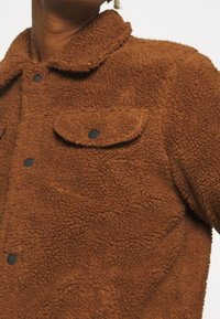 Chaqueta de forro polar marrón con una superficie texturizada, cuello y dos bolsillos frontales asegurados con botones negros.