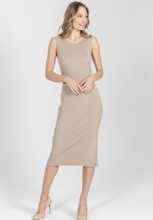 Kobieta uśmiechająca się, ubrana w beżową, bezrękawnikową sukienkę midi z prążkowanego materiału i beżowe szpilki, stojąca na tle jednolitego jasnoszarego tła.