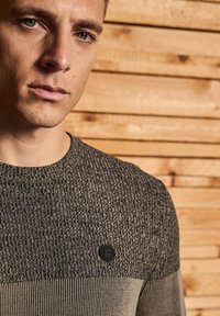 Gestrickter Pullover in Dunkelgrau mit strukturierten und glatten Abschnitten, ausgestattet mit einem runden Hals und einem runden schwarzen Logo-Akzent auf der Brust.