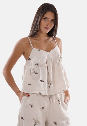 Mujer con largo cabello castaño lleva un top beige de tirantes finos y pantalones cortos a juego con patrones bordados de hojas en negro y marrón.
