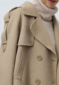 Nais, kellel on seljas beež villane kahekordne jakk heleda roosa soonikkoes turtlenecki sviitri peal, tuues esile jakki detailid ja nupud.