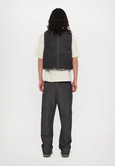 Gilet gris rembourré avec un ourlet arrondi, doté d'une poche arrière, porté par-dessus une chemise beige clair. Accompagné d'un pantalon large gris assorti.