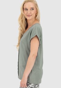 T-shirt verde chiaro a V con maniche corte, realizzata in un tessuto morbido. Presenta un piccolo logo a forma di cuore decorativo all'orlo e una vestibilità comoda.