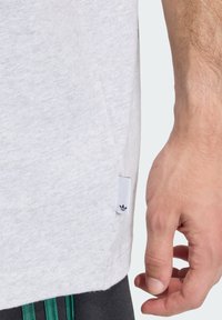 Sivé bavlnené tričko s bočným rázporkom a čiernou nášivkou loga Adidas na leme, ktoré prezentuje mäkký, textúrovaný povrch. Ruka tesne mimo rámu.