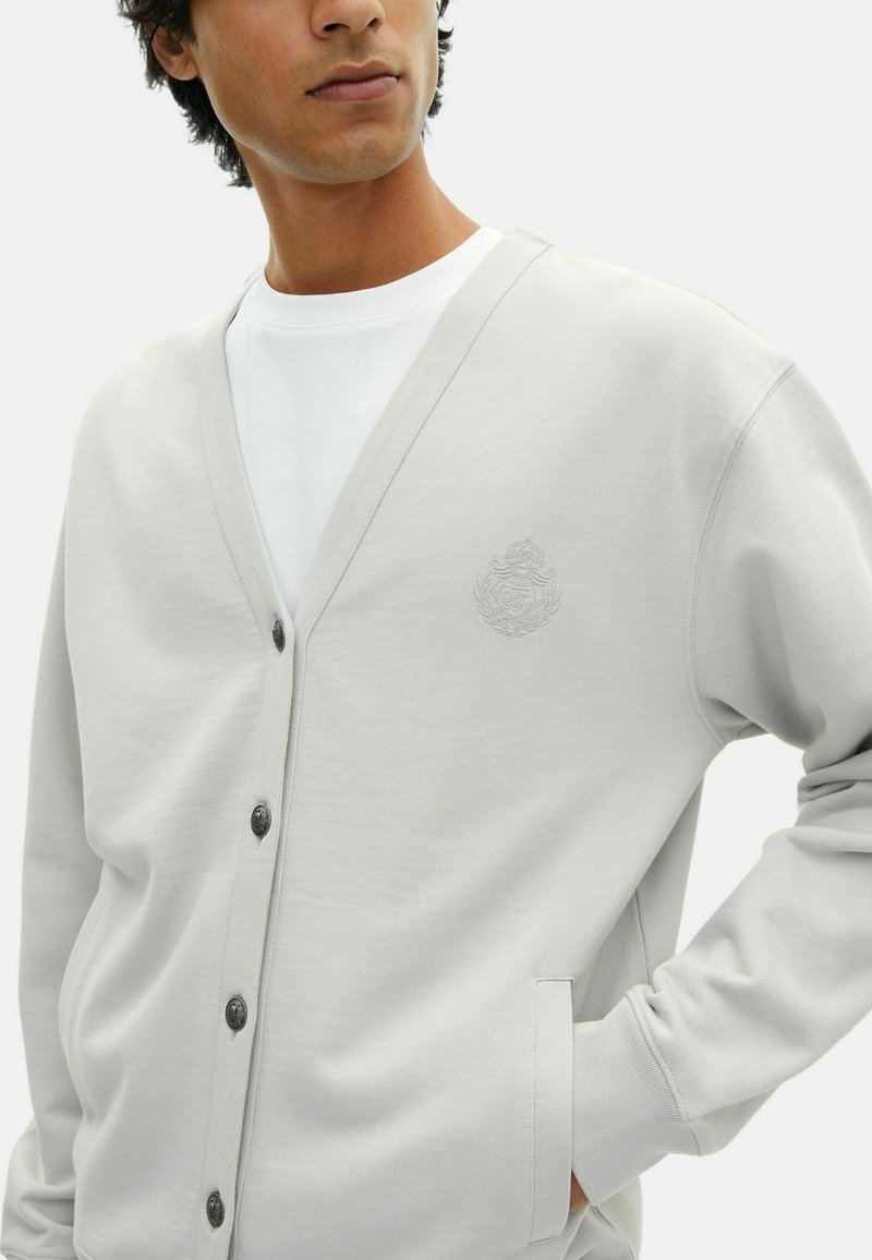 Mann in einem hellgrauen, geknöpften Cardigan über einem weißen T-Shirt, mit der Hand in der Tasche, und einem dezenten gestickten Wappen auf der linken Brust.
